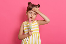 Charming Girl Holding Ice Cream And Touching Her Forehead, Has Headache And High Temperature, Child Wearing Summer Dress, Posing Isolated Over Pink Background.