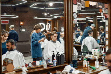 Serving client. Professional barber girl, female hairdresser working with hair clipper, serving young handsome guy sitting in barber shop chair