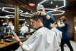 © Svitlana - Side view of a young guy sitting in barber shop chair and using his smartphone while professional barber making trendy haircut for him