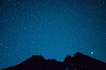 Stars dot the night sky above the silhouette of a broken volcanic mountain peak.