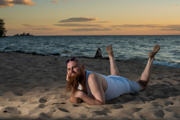 Wall Mural - Funny bald man with red beard posing on the beach at sunset. A humorous male parody of a glamorous girl.