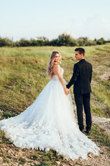 Full length body portrait of young bride and groom enjoying romantic moments outside at sunset in beautiful summer day. Wedding couple. Standing face to face with the green hills on background.