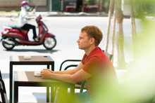 Portrait Of Smile Handsome Young Blond Man With Mole In Casual Style Red T-shirt Sit In Modern Cafe, Happy Look Away, Waiting Someone Or Met Already. Human Relationship, Good Future Changes Concept