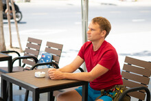 Side Portrait Of Smile Handsome Young Blond Man With Mole In Casual Red T-shirt Sit In Modern Cafe, Happy Look Away, Waiting Someone Or Met Already. Human Relationship, Good Future Changes Concept
