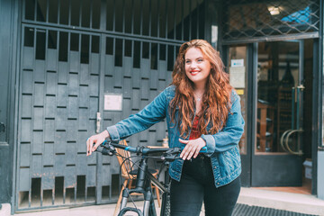 Wall Mural - Fashion portrait of smiling Red curled long hair caucasian teen girl on the city street walking with bicycle. Natural people beauty urban life concept image.