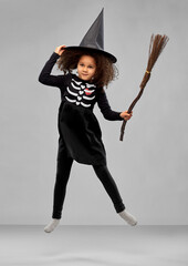 Wall Mural - halloween, holiday and childhood concept - smiling african american girl in black costume dress and witch hat with broom over grey background