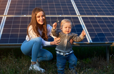 Young mother supporting her little son in making his first steps near solar panels, concept of modern family