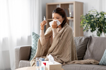 Wall Mural - health, cold and people concept - sad sick young asian woman in blanket with nasal spray medicine and paper tissue at home