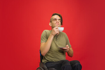 Wall Mural - Joyful coffee, tea time. Caucasian young disabled man's portrait on red studio background. Beautiful model in casual style. Concept of human emotions, facial expression, sales, ad, inclusion