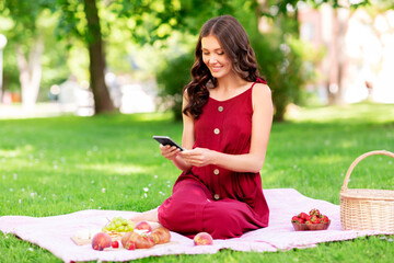 Poster - leisure and people concept - happy smiling woman with smartphone, picnic basket and food sitting on blanket at summer park