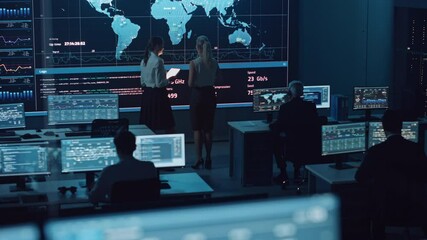 Fototapete - Two Software Engineers Talking Next to a Big Screen in a Modern Monitoring Office with Live Analysis Feed with Charts. Monitoring Room Big Data Scientists and Managers Sit in Front of Computers.