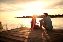 Young Couple Sitting On Wooden Platform By The Lake, Looking Each Other, Talking At Sunset