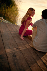 Wall Mural - man and woman sitting on the wooden platform at sunset by the river, enjoying