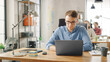 © Gorodenkoff - Handsome Young Man in Glasses and Shirt is Working on a Laptop in a Creative Business Agency. They Work in Loft Office. Diverse People Working in the Background. He's in Good Mood.