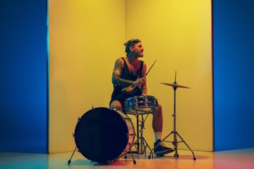 Rockstar. Young musician with drums performing on yellow background in neon light. Concept of music, hobby, festival, entertainment, emotions. Joyful, inspired drummer. Colorful portrait of artist.