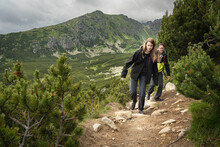 Two People Hiking On Rocky Trail In HIgh Tatras Mountains In Slovakia Surrounded By Dwarf Pine And Other Coniferous Trees. Top Of Mountain In Clouds In Background