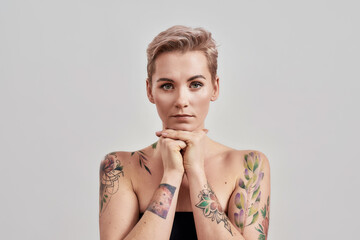 Portrait of half naked tattooed woman with short hair looking at camera, holding chin on hands isolated over light background