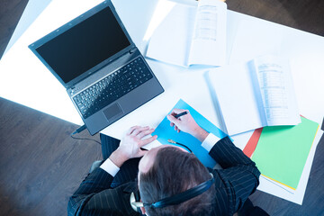 Poster - Top view of a businessman with a laptop and documents. The person with headphones is in the office. Telecommuting. Office work. The employee checks the documents.