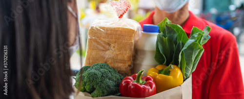 Panoramic banner. food delivery service man wearing protection face mask handing fresh food set bag to young woman customer receiving order from courier at home, food delivery, online shopping concept