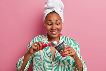 Sticker - Positive excited dark skinned young woman has temptation to eat sweet delicious cold ice cream, holds special spoon, smiles happily, wears domestic robe and bath towel on head, stands indoor