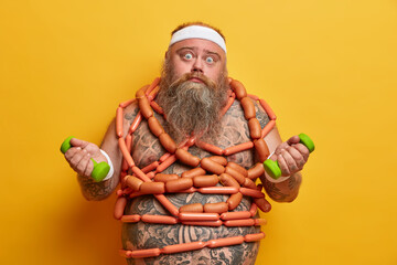 Wall Mural - Problem of obesity. Shocked tired man with big tummy and thick beard, gets lots of energy from sausages, trains muscles, holds dumbbells, concerns about health, isolated on yellow background