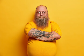 Handsome bearded man keeps arms folded, looks thoughtfully away, has plump body, dressed in casual clothes, makes up plan how to lose weight, isolated over yellow background. Pensive indecisive guy