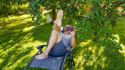 Middle-aged man with mobile phone in his hands lies on a deck chair in the garden under a tree in summer time. Vacation and recreation, technology concept.