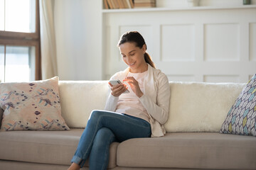 Smiling young attractive woman sitting on cozy couch, enjoying communicating with friends in social network or shopping online application on smartphone, relaxed free leisure weekend pastime concept.