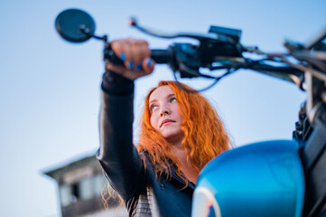 Curly red-haired woman in a black leather jacket sits on a motorcycle. Portrait of a serious girl driving a bike.