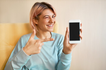 Wall Mural - Indoor shot of beautiful positive young European female sitting on armchair with her new smart phone, winking, pointing fore finger at black display with copy space for your content or information