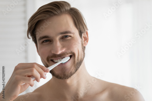 Head shot portrait close up smiling young man brushing teeth, looking at camera, personal oral hygiene concept, satisfied handsome guy enjoying morning routine, standing in bathroom