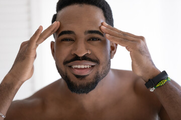 Head shot portrait smiling African American young man with naked shoulders checking wrinkles or acne on forehead, looking at camera, facial cosmetology, treatment, skincare concept