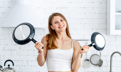 Wall Mural - Funny young housewife with frying pans in modern kitchen laughing. Closeup