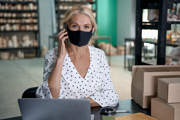 Wall Mural - Female business owner wearing protective face mask talking by phone and working on laptop while sitting in her art studio or craft pottery shop with handmade ceramic product