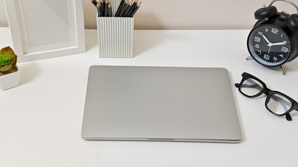 Wall Mural - Close up of minimal workspace with laptop, glasses, clock, pencils, and frame on white table.