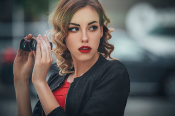 Beautiful brunette sexy spy agent (killer or police) woman in leather jacket and red luxury dress with a binocle in her hand watching after someone in european city