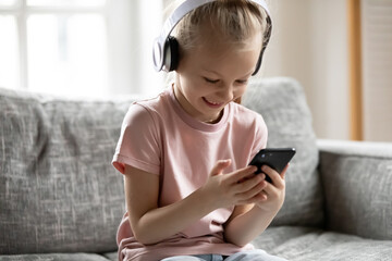 Overjoyed little adorable blonde girl wearing wireless headphones, watching funny video in social network on smartphone or playing online mobile games alone at home, children and technology concept.