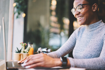 Cheerful afro american web designer satisfied with completing project earning money online while sitting in cafe interior near advertising area,smiling female student receiving good job offer on email