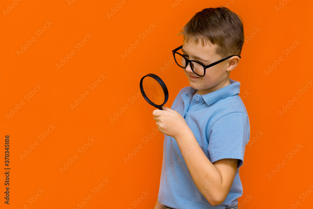 Beautiful european young boy with glasses holding and looking through magnifying glass.Orange wall. - obrazy, fototapety, plakaty 