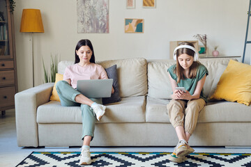 Wall Mural - Young female networking while her daughter in headphones scrolling in smartphone