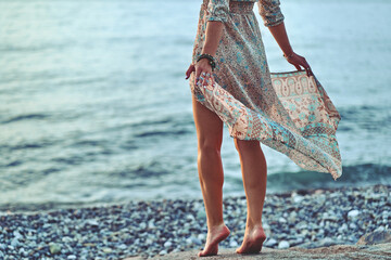 Boho woman with long legs and waving dress standing on a stone by the seashore at sunset