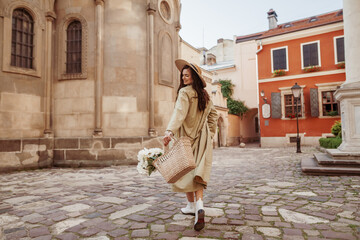 Happy smiling woman wearing straw hat, long trench coat, holding basket with flowers, walking in street of European city. Lifestyle, travel concept. Full-length portrait. Copy, empty space for text