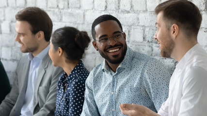 Close up smiling African American businessman wearing glasses chatting, diverse candidates applicants interns talking while waiting for job interview, sitting in queue in row, recruitment concept