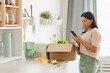 © Seventyfour - Waist up portrait of modern mixed-race woman holding smartphone while standing by box of food in kitchen, mood delivery service and mobile app concept