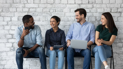 Wall Mural - Young overjoyed diverse candidates business people laughing at joke, chatting, using electronic mobile devices, waiting for job interview or exam, sitting on chairs in queue, employment concept