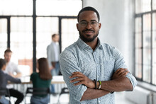 Head Shot Portrait Confident Young African American Businessman Wearing Glasses Looking At Camera, Successful Executive Startup Founder Standing In Modern Office Room With Arms Crossed