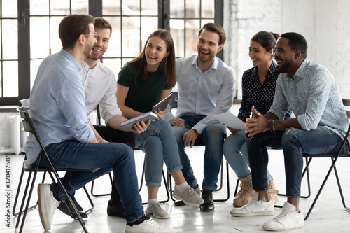 Happy diverse colleagues laughing at joke, having fun at briefing, sitting on chairs in modern office, sharing ideas, positive overjoyed employees staff talking, chatting, good relationship