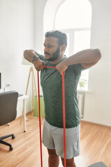 Wall Mural - For body tone. Young active man looking away, exercising with resistance band during morning workout at home. Sport, healthy lifestyle