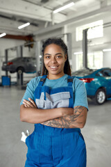 Wall Mural - Here to serve you. Portrait of young african american woman, professional female mechanic in uniform smiling at camera, standing in auto repair shop. Car service, repair, maintenance, people concept