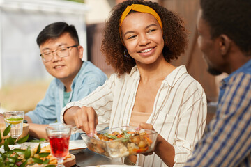 Wall Mural - Portrait of smiling African-American woman sharing food while enjoying dinner with friends and family outdoors at Summer party, copy space